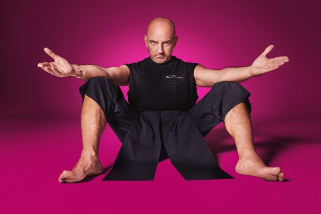 Homem careca sentado de braços abertos sobre fundo magenta.