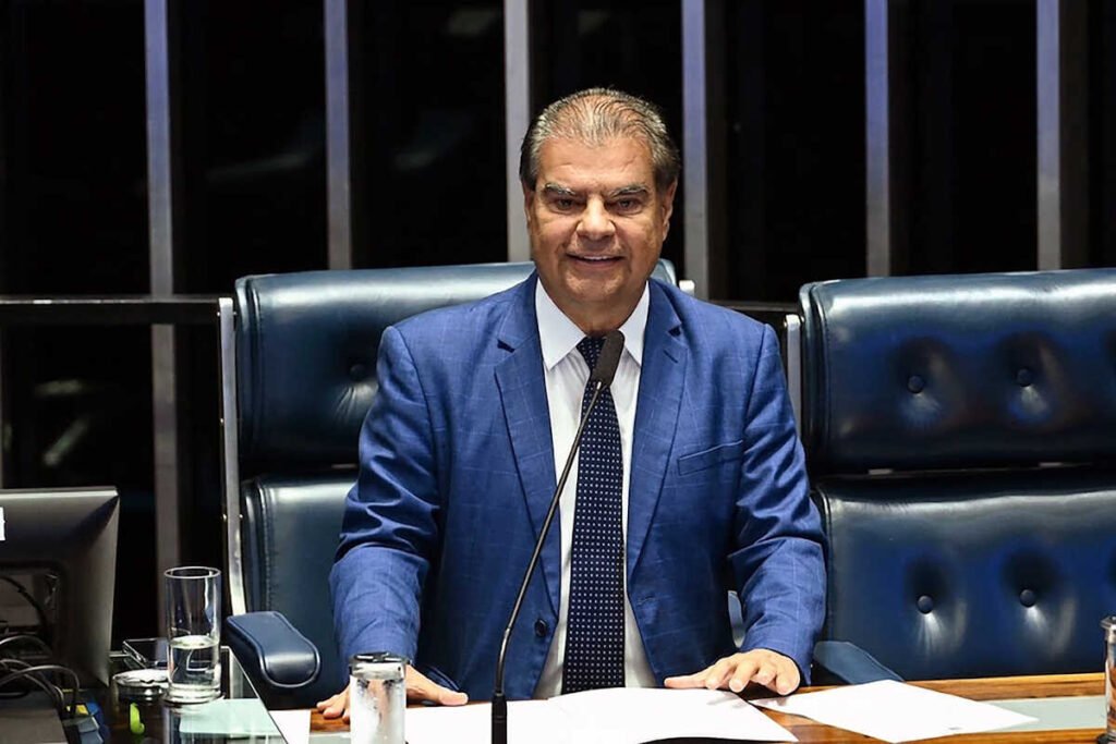 Homem em terno azul sentado em plenário legislativo.