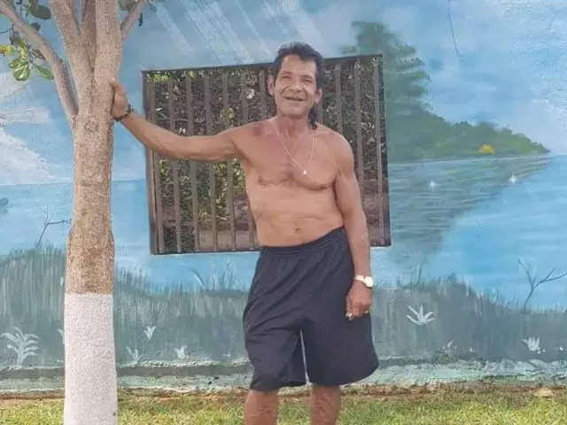 Homem sorridente sem camisa encostado em uma árvore pequena.
