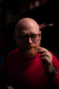 Homem careca com barba ruiva e óculos usando blusa vermelha.