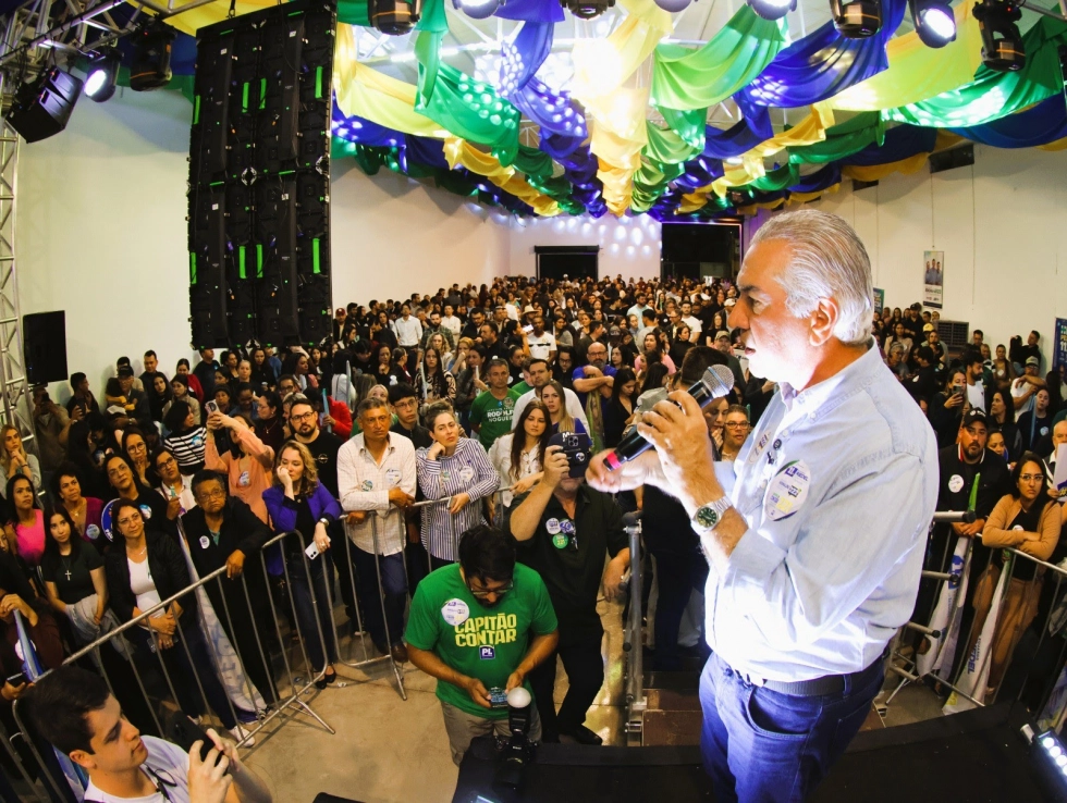 Homem discursando para grande multidão em evento com cores do Brasil.