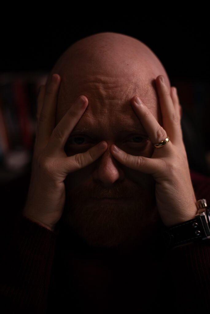 Homem careca com mãos no rosto em iluminação dramática.