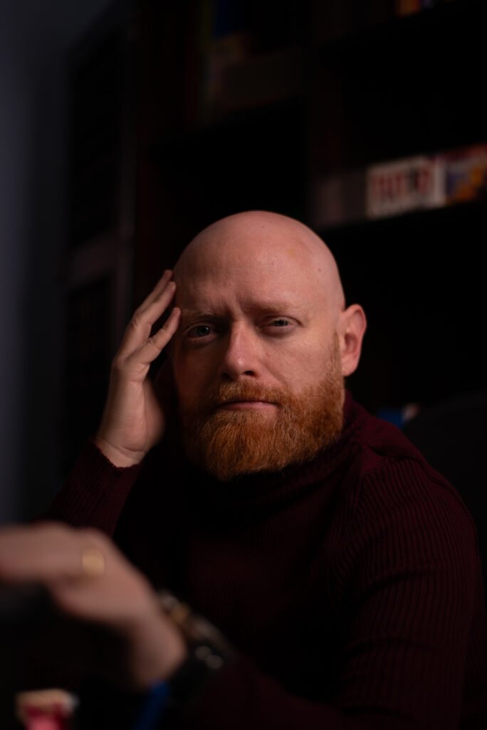 Homem careca de barba ruiva usando gola alta bordô.