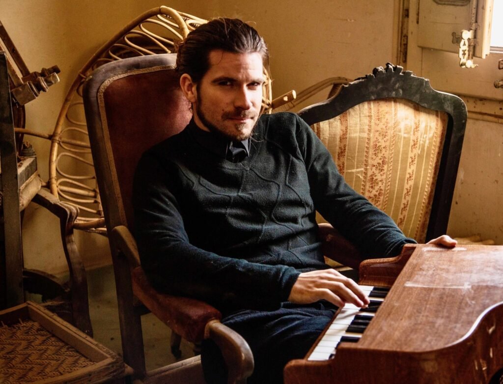 Homem sentado em poltrona antiga tocando pequeno piano de madeira.