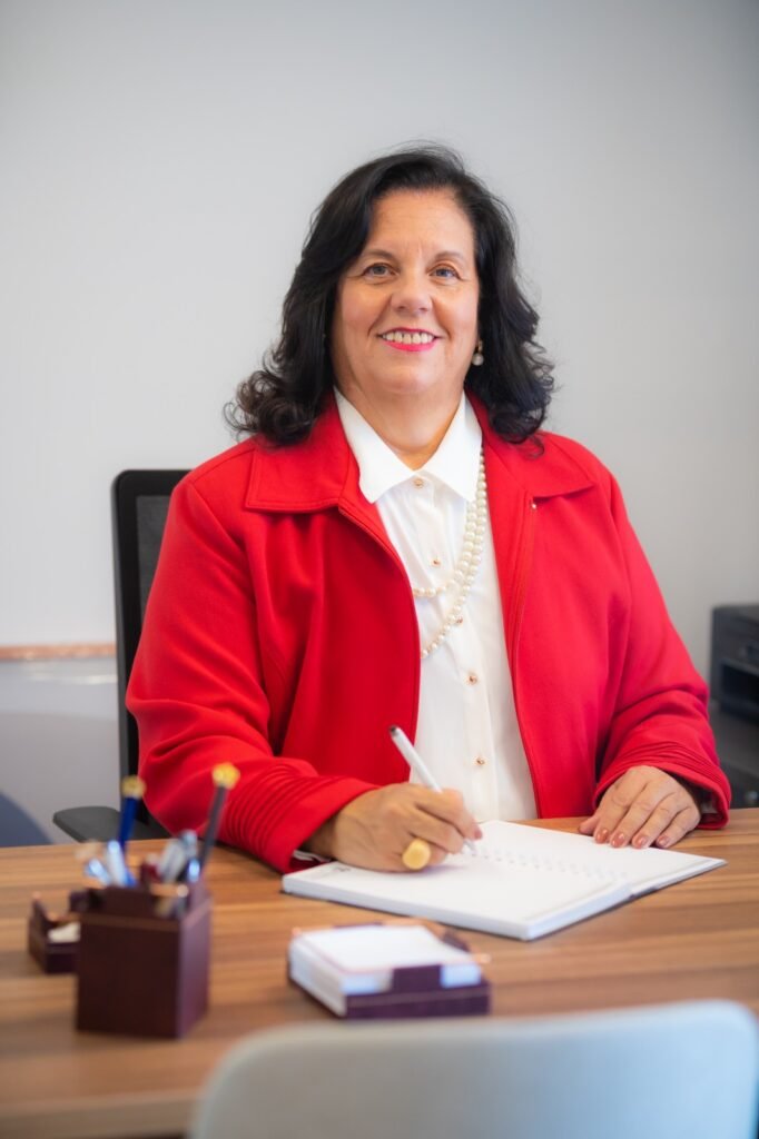 Mulher de blazer vermelho sorrindo ao escrever em caderno.