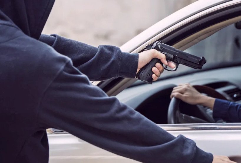 Pessoa encapuzada apontando arma para motorista dentro de um carro.