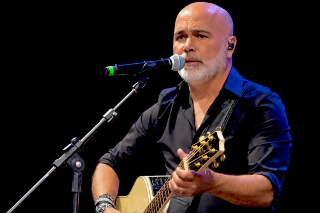 Homem careca com barba grisalha cantando e tocando violão.