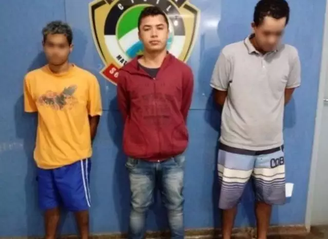 Três homens detidos em frente a um escudo policial.