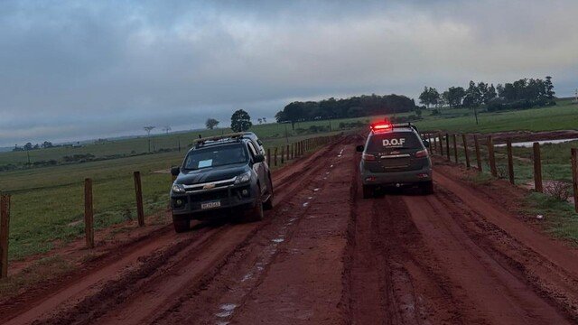 Viatura policial e caminhonete em estrada de terra lamacenta.