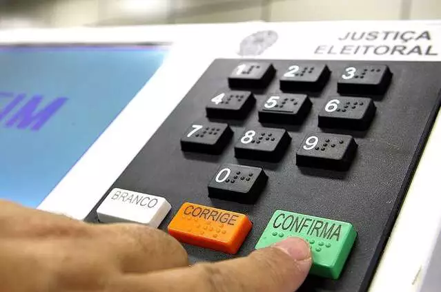 Dedo pressionando o botão Confirma de uma urna eletrônica brasileira.