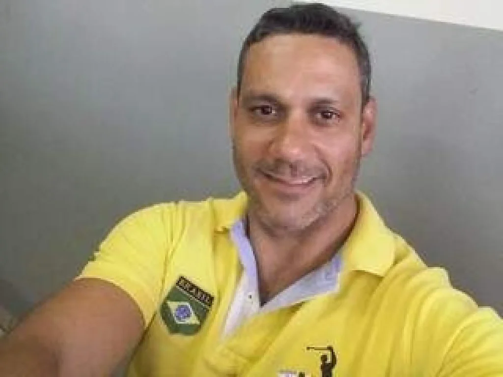 Homem sorridente vestindo camisa polo amarela com bandeira do Brasil.