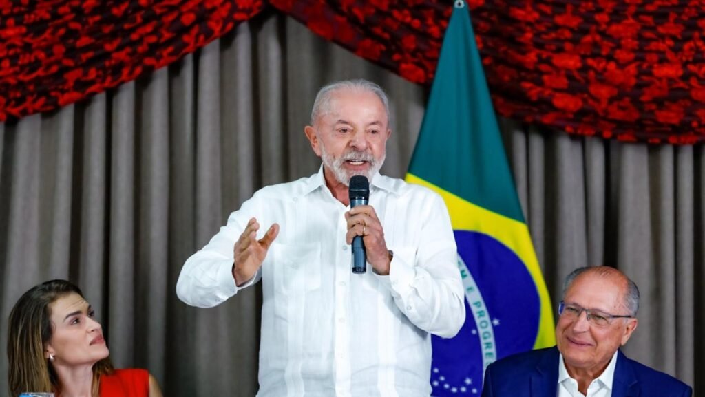 Lula discursa com microfone ao lado de Alckmin e bandeira.