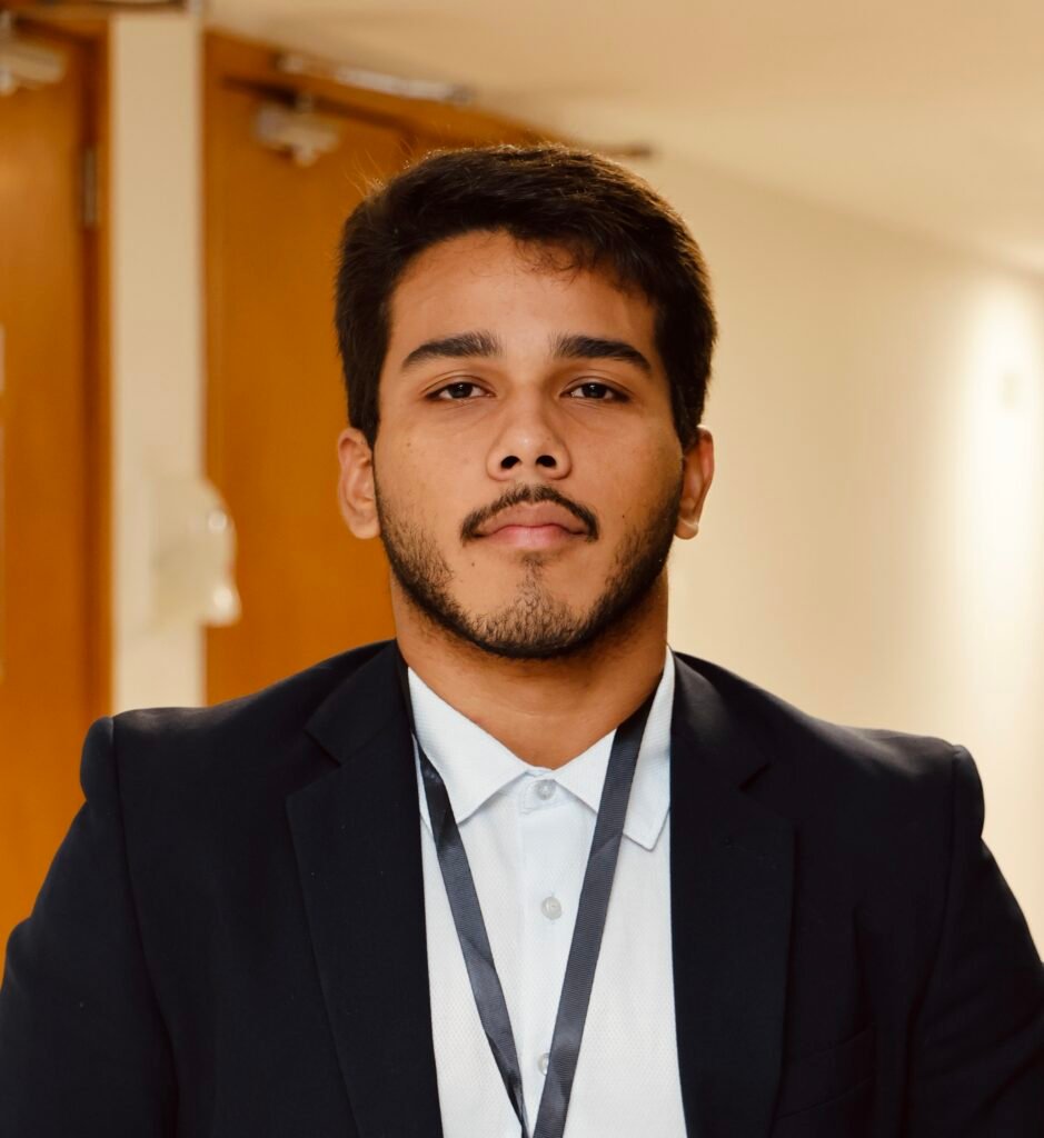 Homem jovem de terno e crachá em ambiente interno.