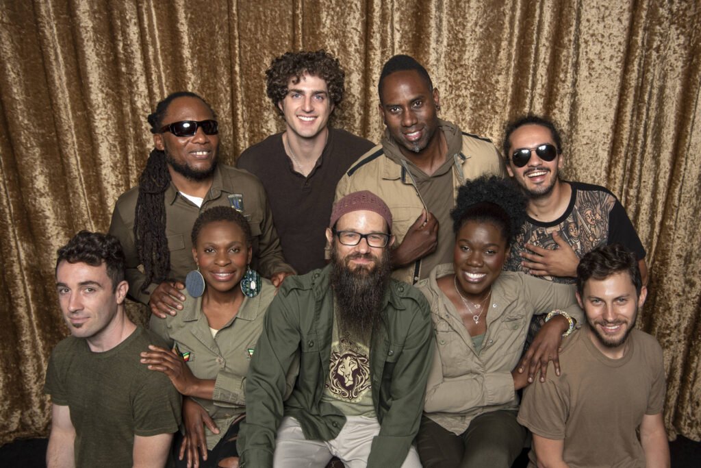Grupo diversificado de nove pessoas posando diante de cortina dourada.