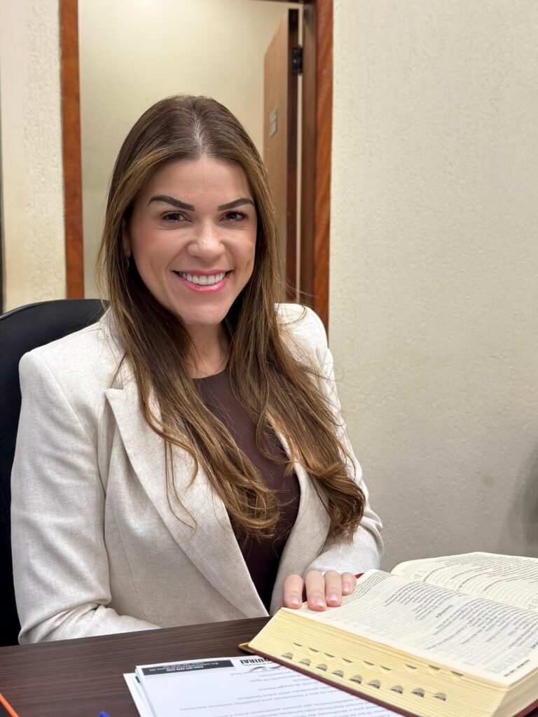 Mulher sorridente de blazer branco sentada à mesa com livro.