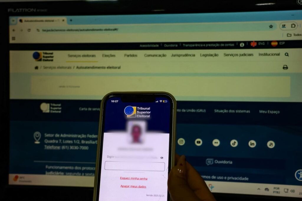 Mão segurando celular com app do TSE diante de monitor.