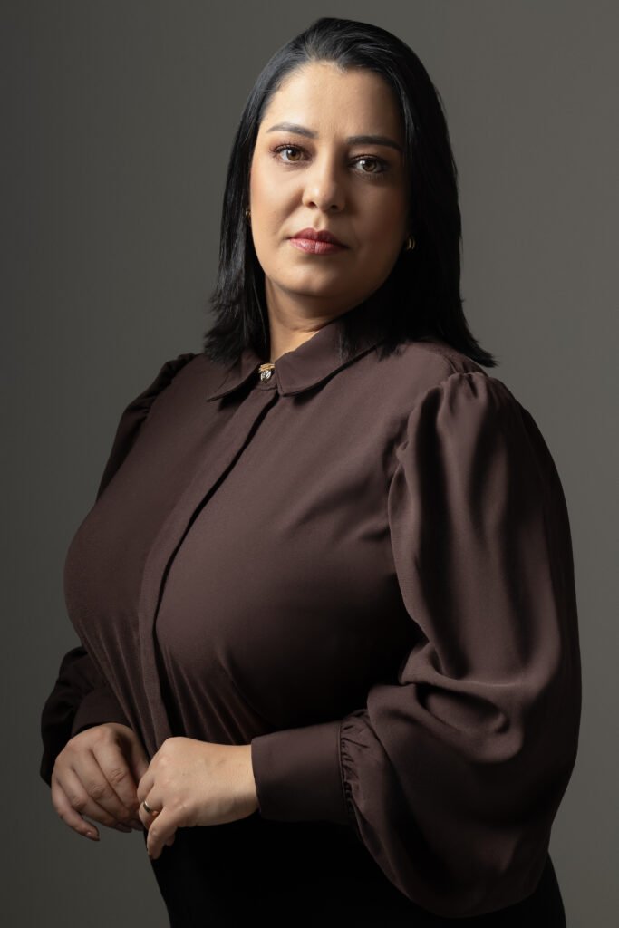 Mulher de cabelos escuros usando blusa marrom em fundo cinza.