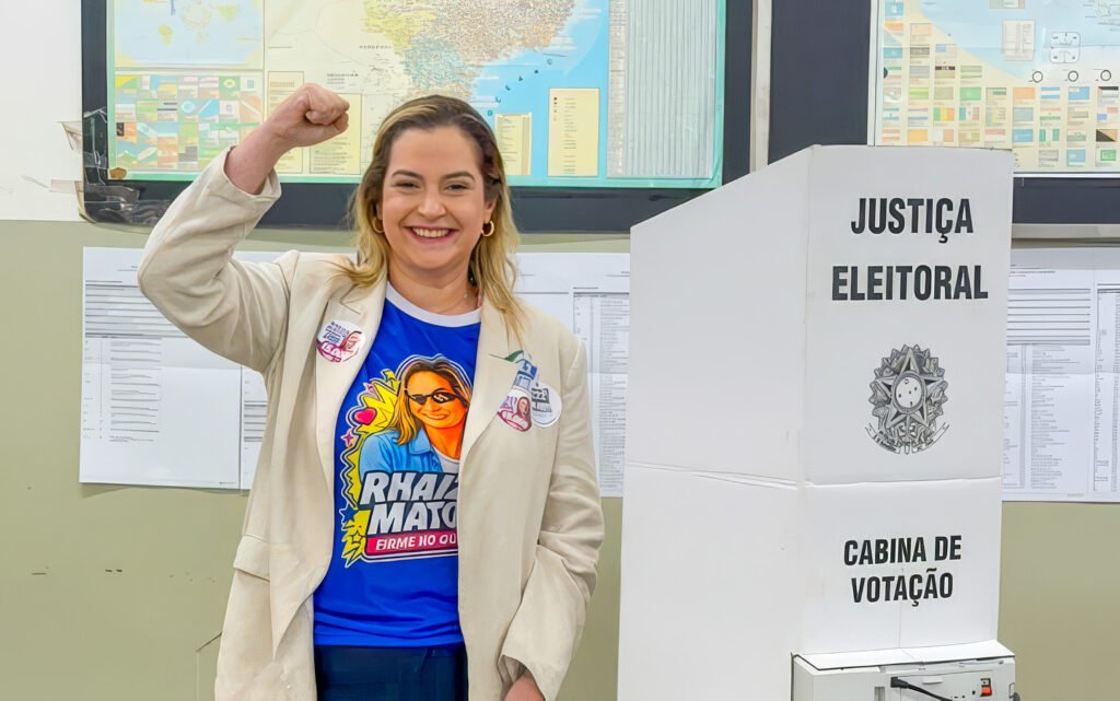 Mulher sorridente com punho erguido ao lado de cabine eleitoral.
