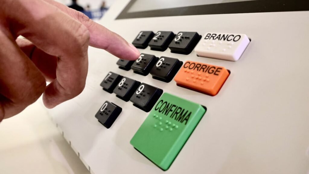Mão pressionando tecla numérica em uma urna eletrônica brasileira.