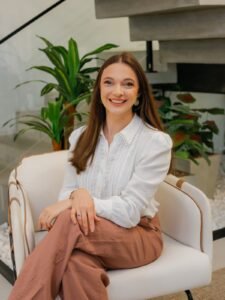 Mulher sorridente sentada em poltrona branca com plantas ao fundo.