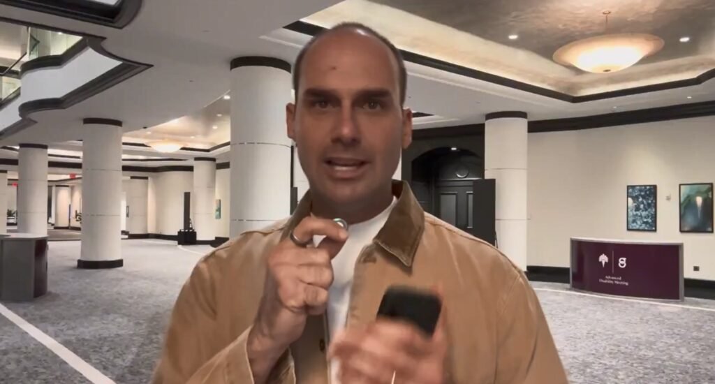 Eduardo Bolsonaro fala em saguão segurando microfone e celular.