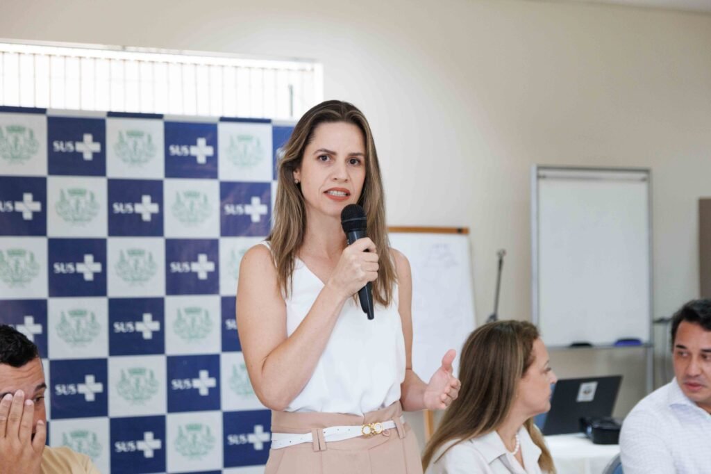 Mulher discursando ao microfone em frente a painel do SUS.