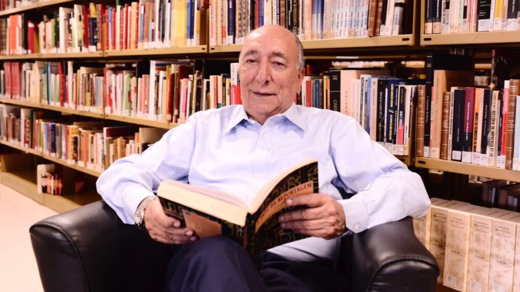 Homem idoso lendo um livro em frente a uma estante.