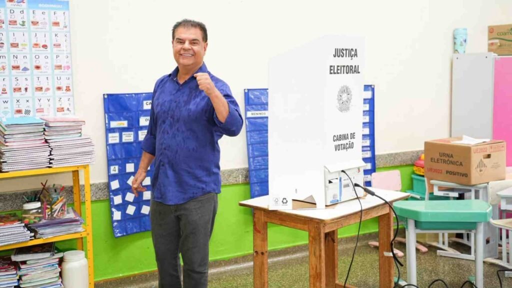 Homem sorridente em seção eleitoral ao lado de cabine de votação.