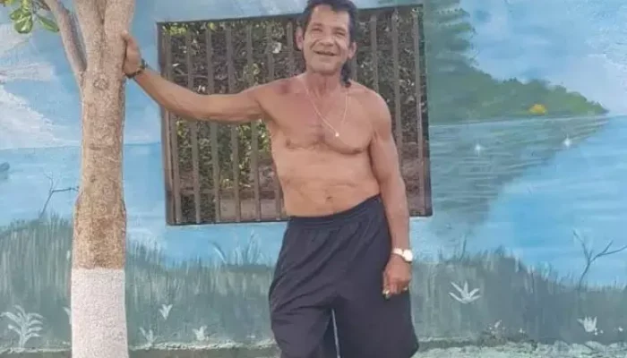 Homem sorridente sem camisa encostado em uma árvore pequena.