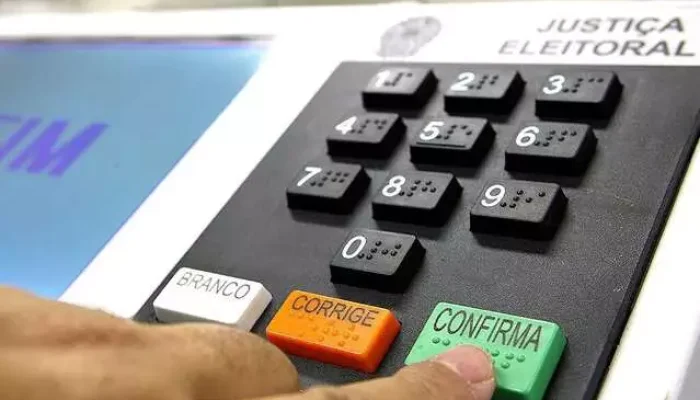 Dedo pressionando o botão Confirma de uma urna eletrônica brasileira.