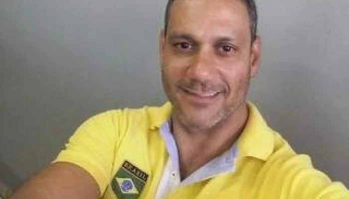 Homem sorridente vestindo camisa polo amarela com bandeira do Brasil.