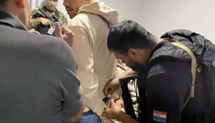 Policiais algemando um homem com boné azul em ambiente interno.