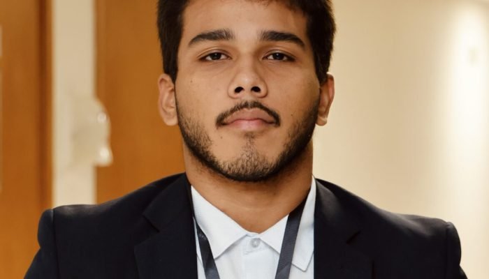 Homem jovem de terno e crachá em ambiente interno.