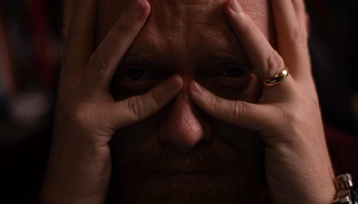 Homem careca com mãos no rosto em iluminação dramática.