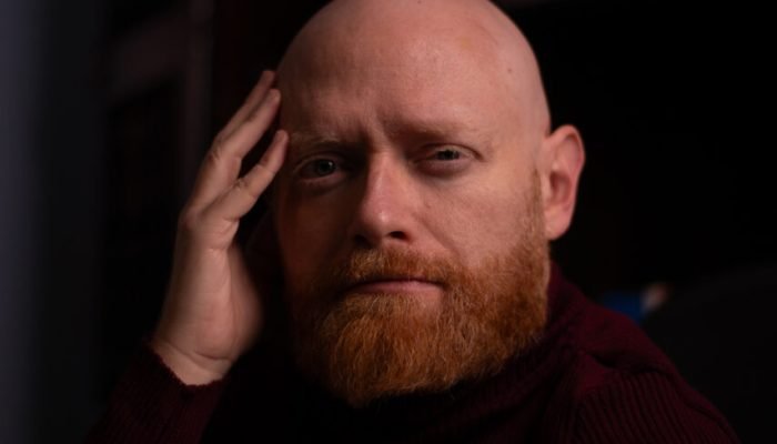 Homem careca de barba ruiva usando gola alta bordô.