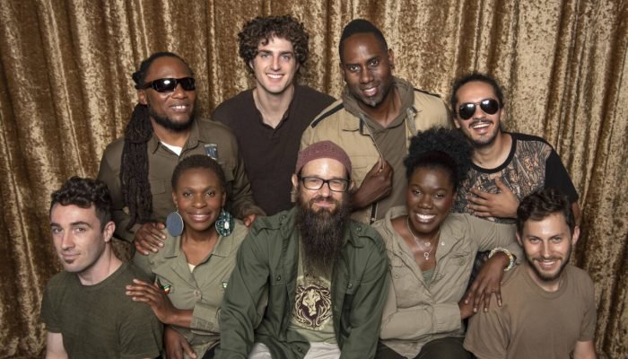 Grupo diversificado de nove pessoas posando diante de cortina dourada.
