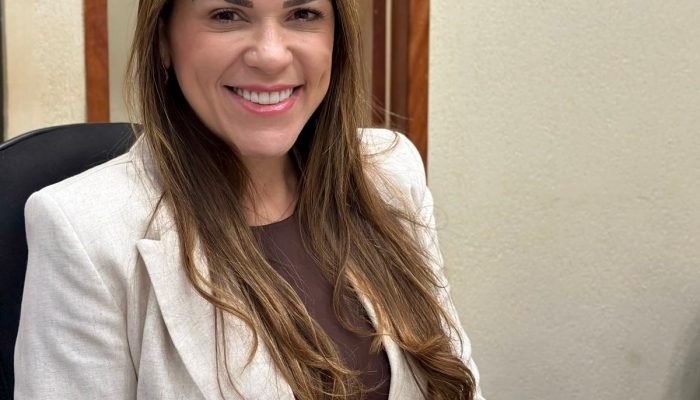 Mulher sorridente de blazer branco sentada à mesa com livro.