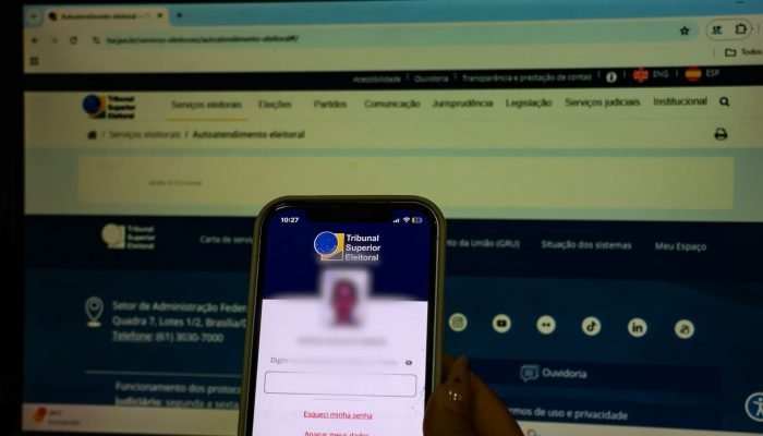 Mão segurando celular com app do TSE diante de monitor.