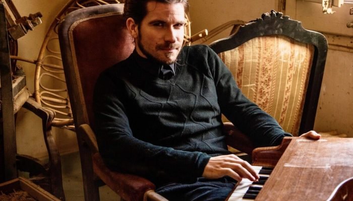 Homem sentado em poltrona antiga tocando pequeno piano de madeira.