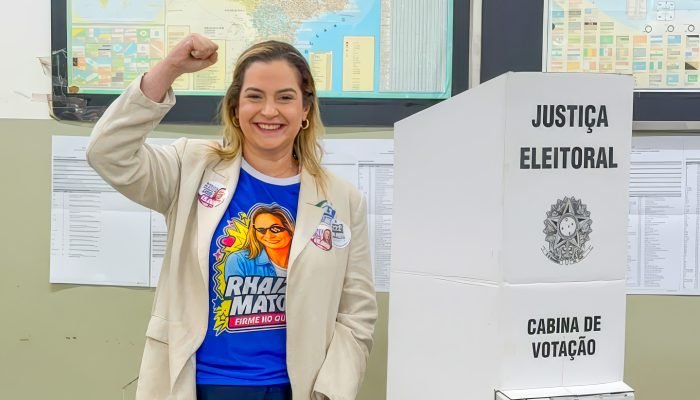 Mulher sorridente com punho erguido ao lado de cabine eleitoral.
