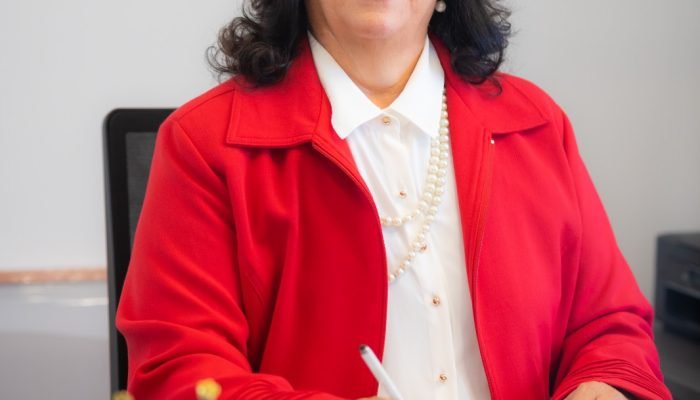 Mulher de blazer vermelho sorrindo ao escrever em caderno.