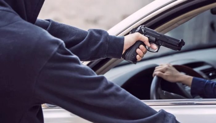 Pessoa encapuzada apontando arma para motorista dentro de um carro.