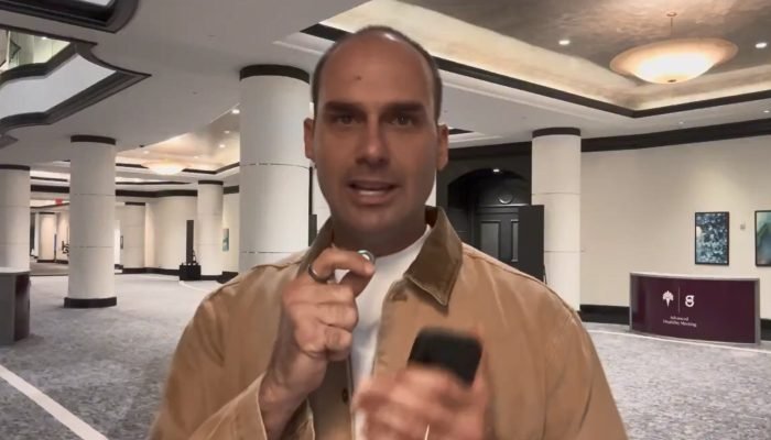 Eduardo Bolsonaro fala em saguão segurando microfone e celular.