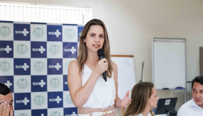 Mulher discursando ao microfone em frente a painel do SUS.