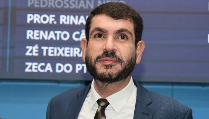 Homem de terno e barba em frente a painel eletrônico.