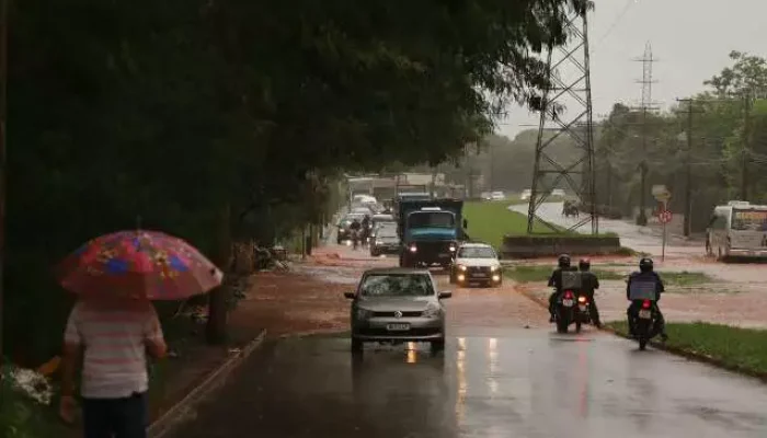 Trânsito em rua alagada sob chuva com pedestre e veículos.