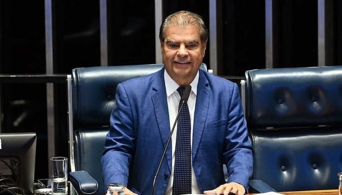 Homem em terno azul sentado em plenário legislativo.