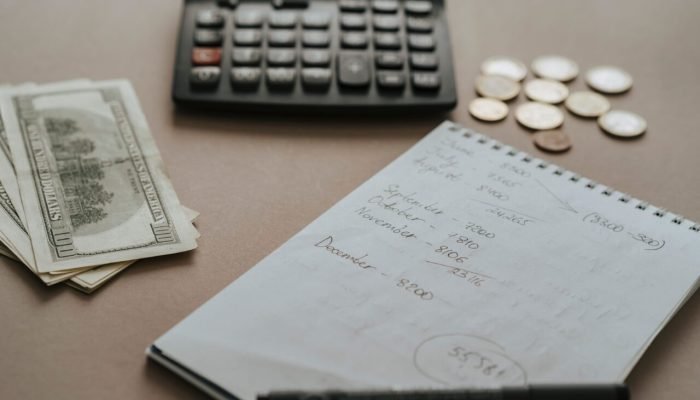 Calculadora, dinheiro, moedas e caderno com anotações financeiras sobre mesa.