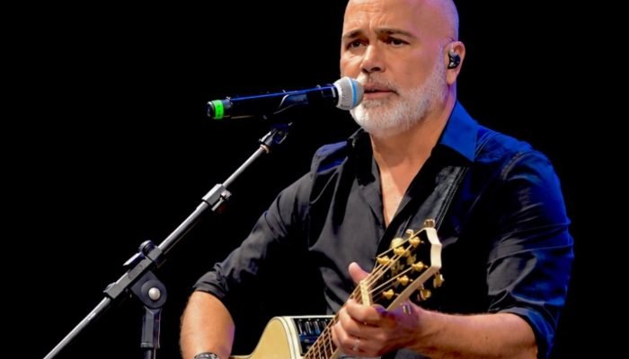Homem careca com barba grisalha cantando e tocando violão.